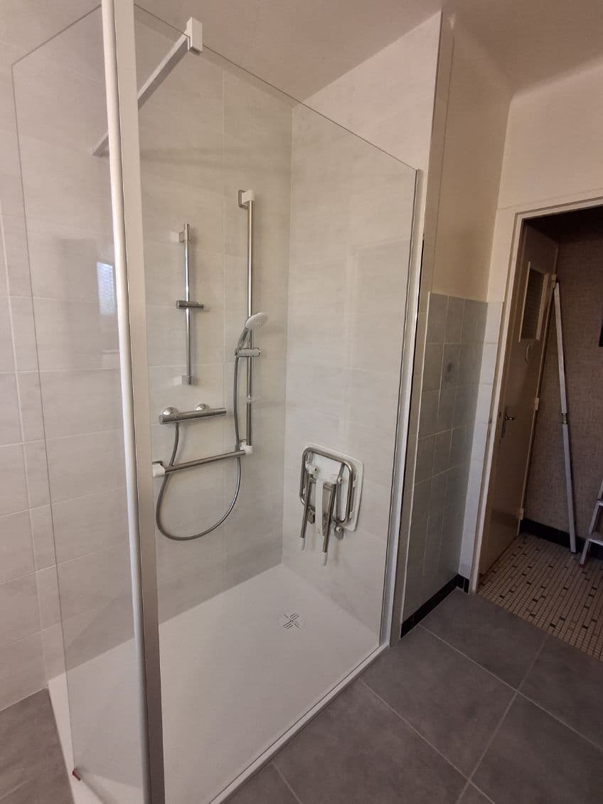 Espace douche adaptée avec une robinetterie anti-brûlure, un siège de douche rabattable et une barre de maintien 